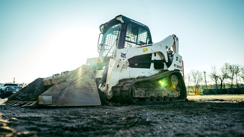 A skid steer loader on a construction site with bright sunlight in the background.